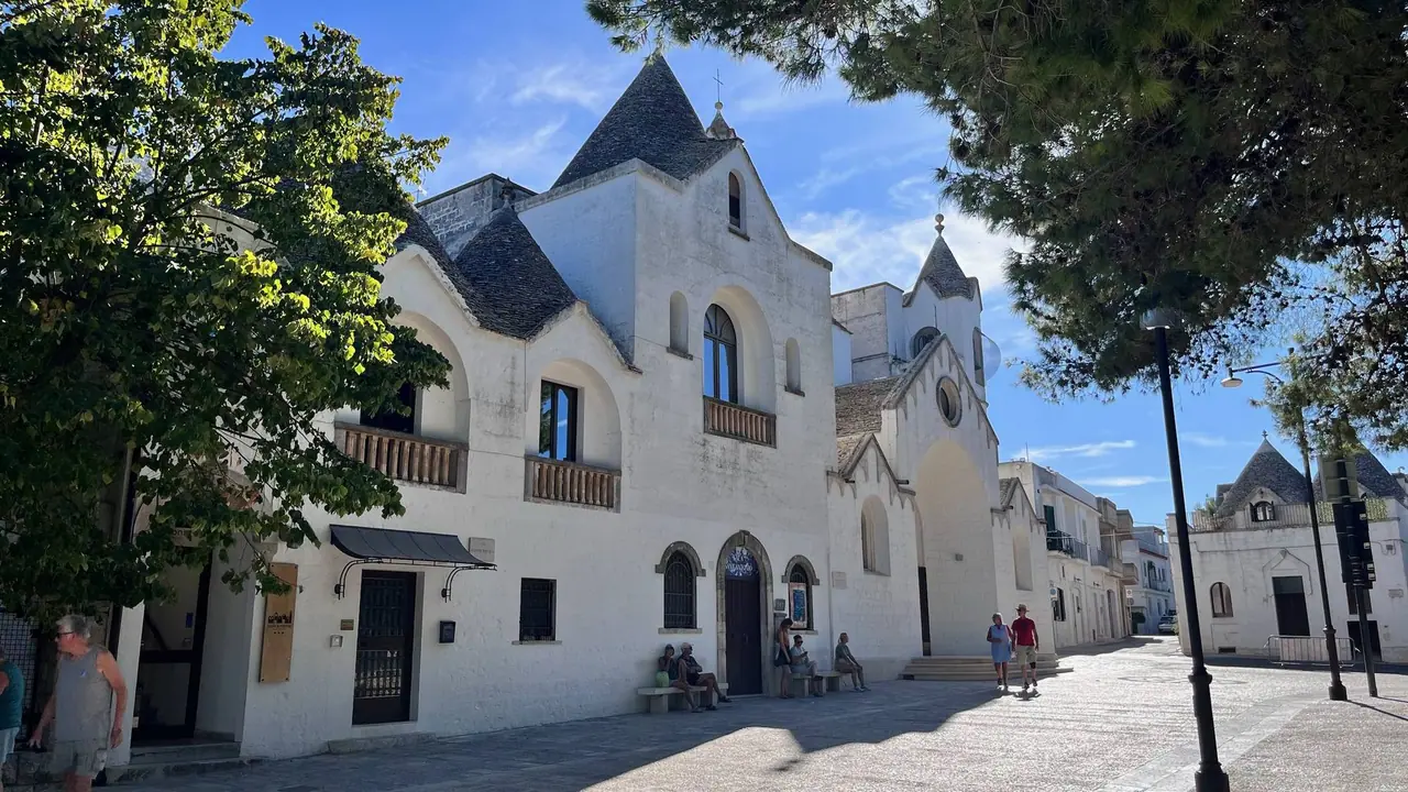 Bílý kostel svatých Cosma a Damiána v Alberobello s kuželovitými věžemi, před kterým si odpočívají turisté na lavičkách pod stromy