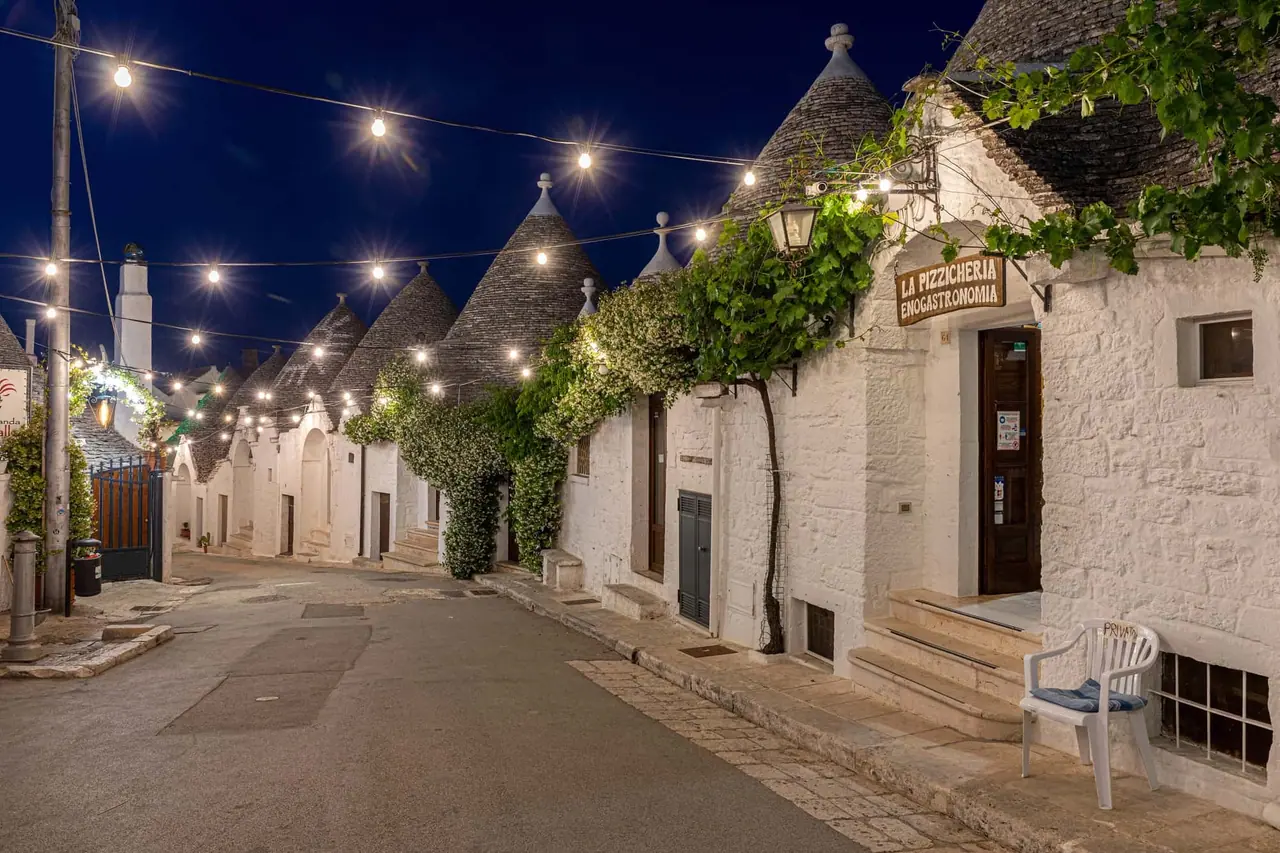 Večerní pohled na typické trulli domky v Alberobello s kuželovými střechami, řetězem světel nad dlážděnou ulicí a pizzerií v bílém kamenném domě