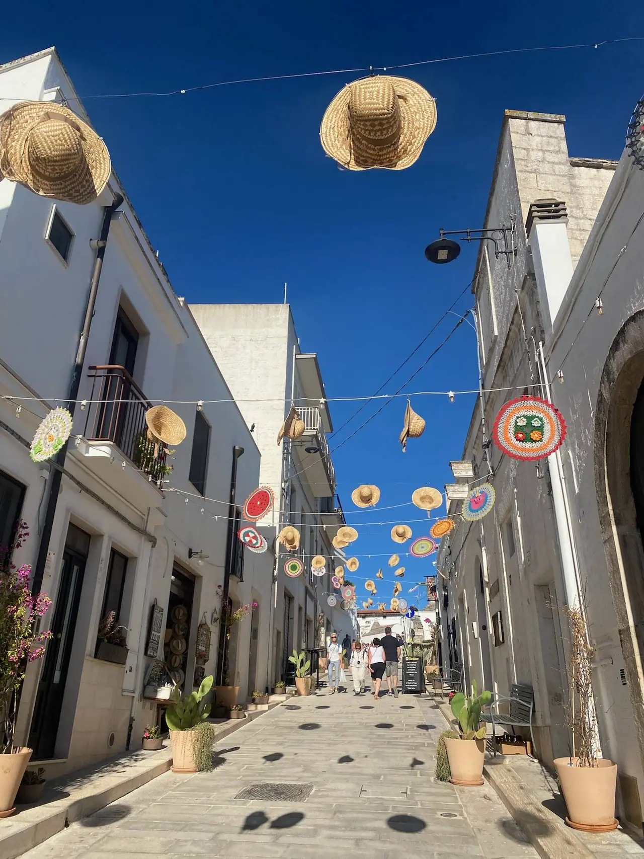 Úzká ulička v Alberobello s bílými domky a barevnými slámovými klobouky visuícími nad hlavami turistů pod modrým nebem