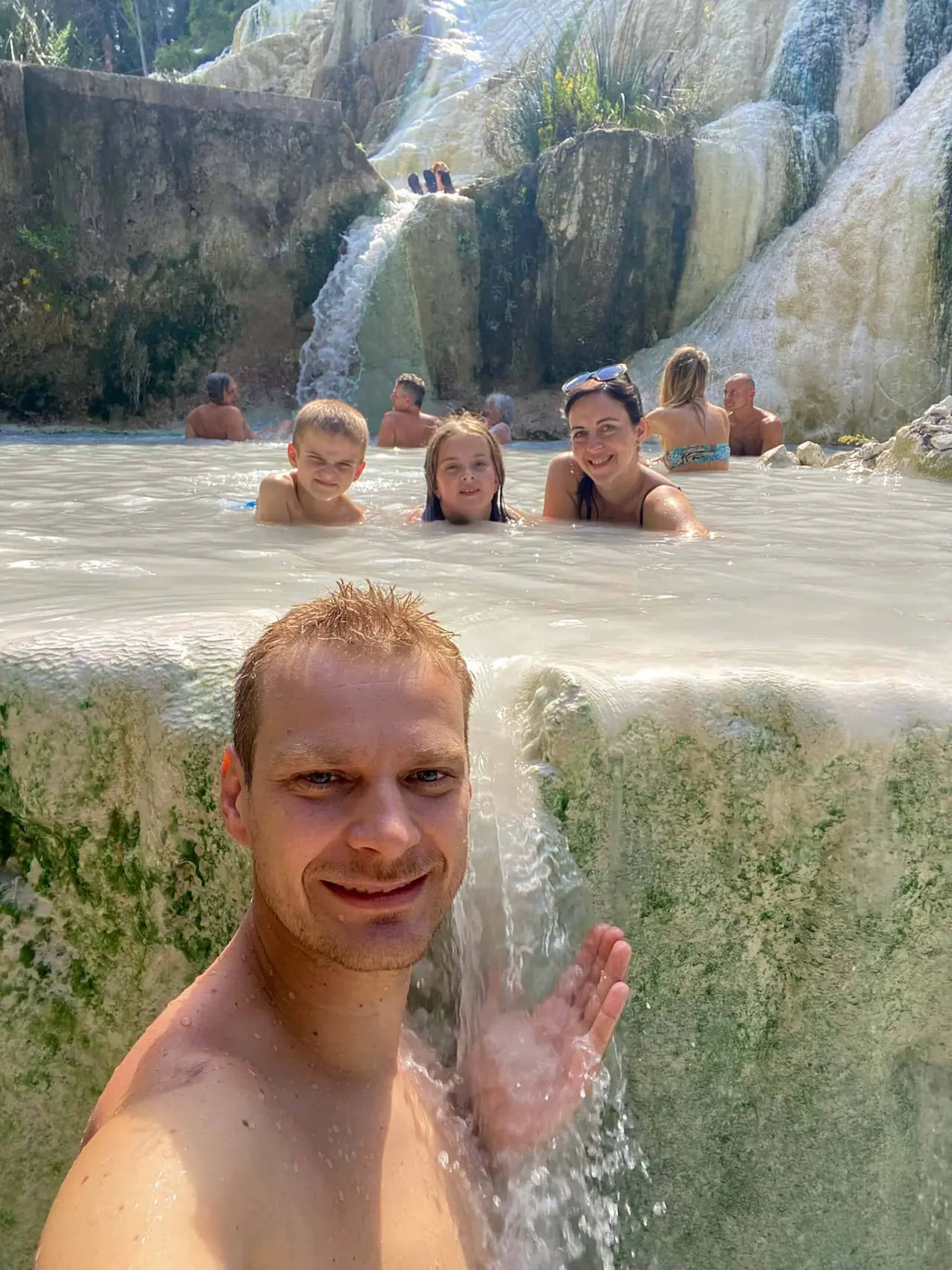 Moje selfie tyčí u bílých vápencových teras v Bagni San Filippo, za ním rodina relaxuje v teplých pramenech