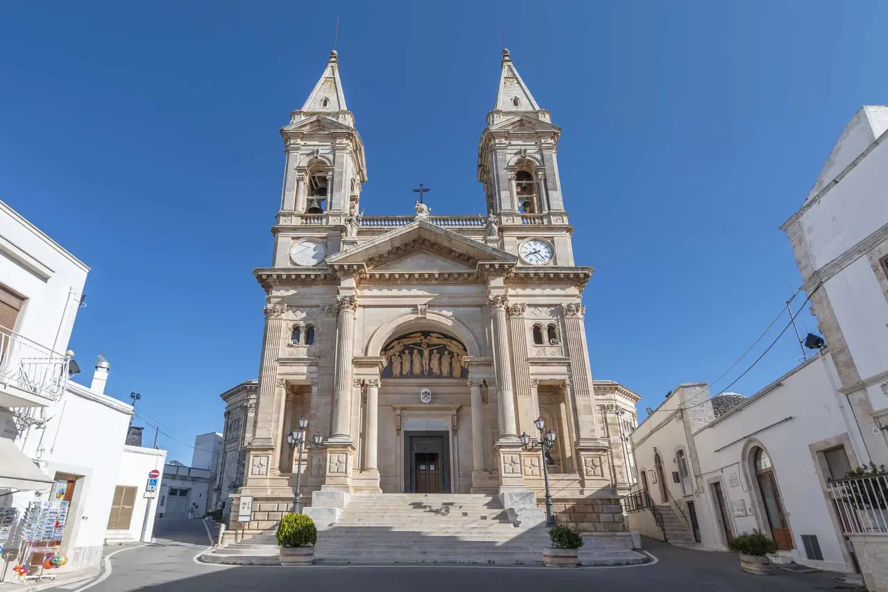 Impozantní barokní bazilika Saints Cosmas and Damiano s dvěma vysokými věžemi a hodinami, obklopená bílými domy v Alberobello