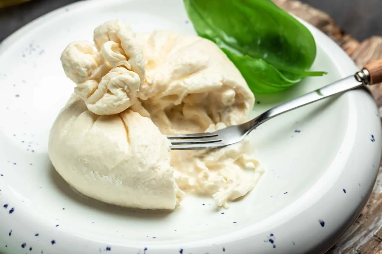 Čerstvá burrata na bílém talíři s vidličkou, sýr rozříznutý do půlky odhaluje krémovou náplň uvnitř