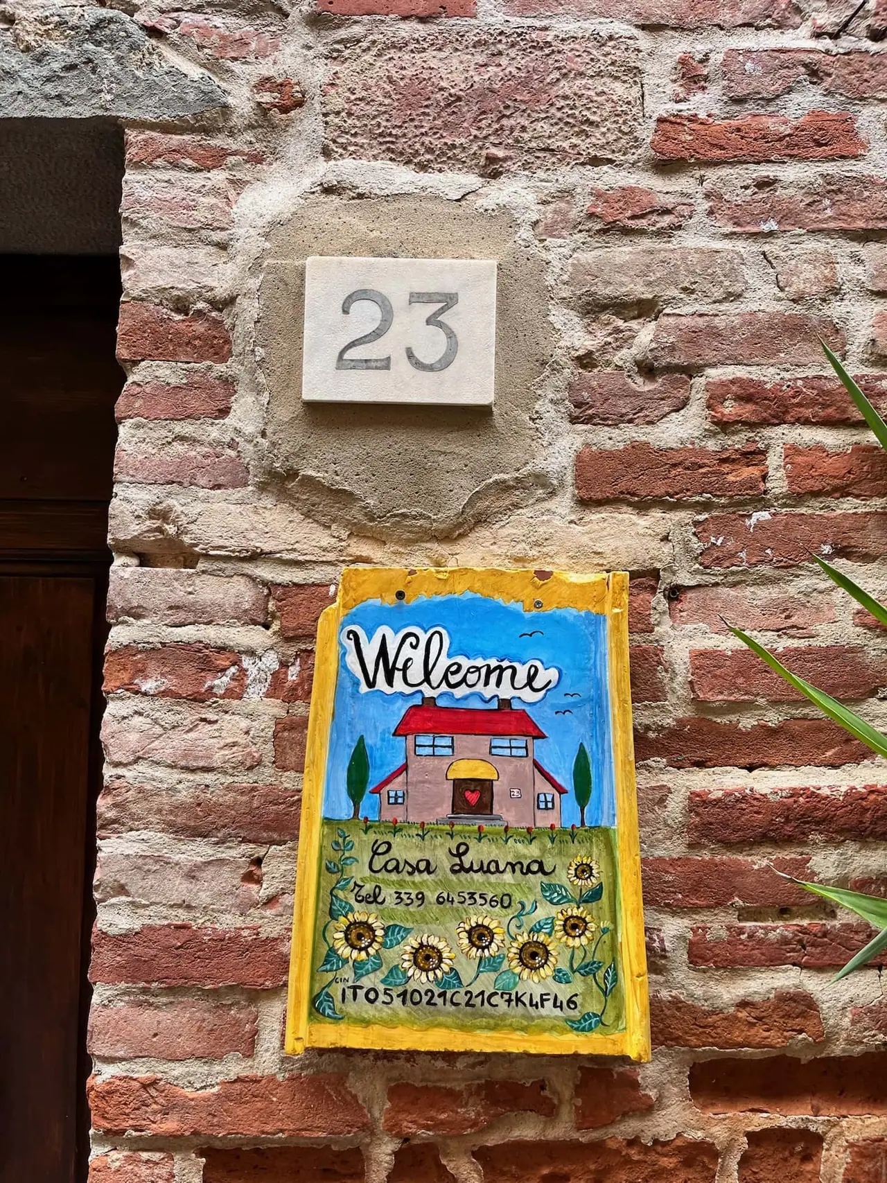 Barevná cedule Casa Luana s malovaným domečkem a slunečnicemi na cihlové zdi domu číslo 23 v Lucignanu