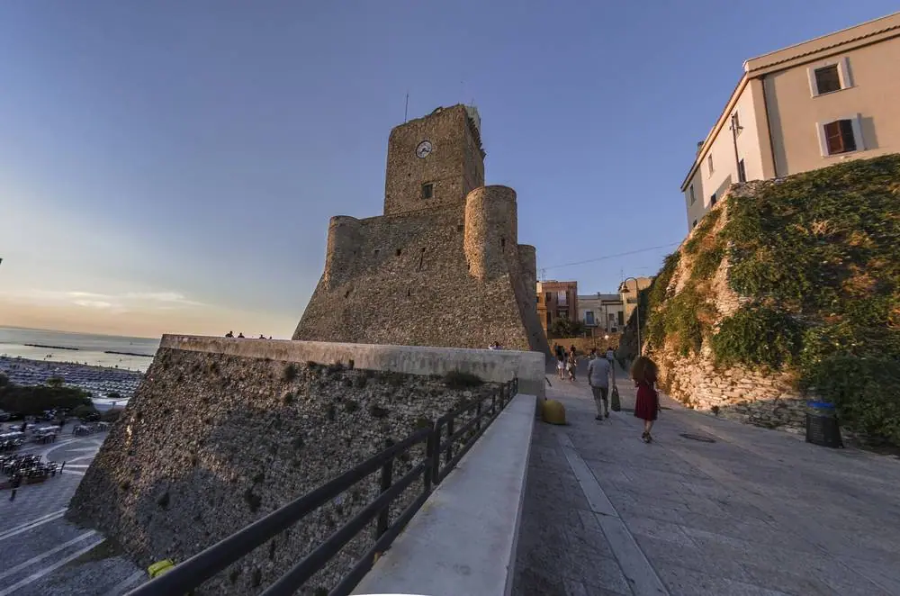 Středověký hrad Castello Svevo v Termoli při západu slunce, kde kolemjdoucí kráčejí po kamenné promenádě podél mohutných hradeb s hodinovou věží.