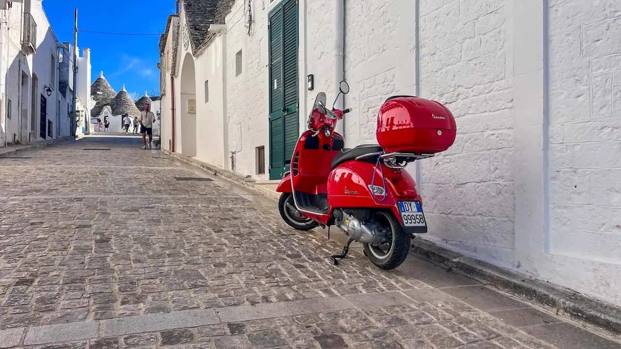 Červená italská Vespa zaparkovaná v úzké dlážděné uličce mezi bílými trulli domečky v historickém centru Alberobella