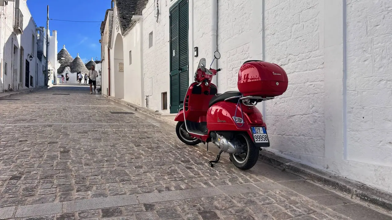 Červená motorka Vespa zaparkovaná v úzké dlážděné uličce mezi bílými domy v historickém centru Alberobello s trulli v pozadí