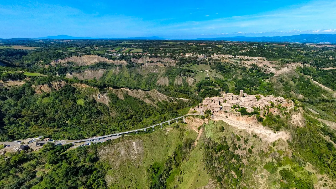 Letecký panoramatický pohled na Civita di Bagnoregio s okolní erozní krajinou calanchi a přístupovým mostem.