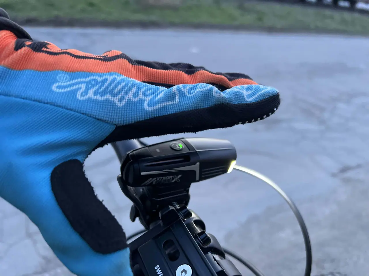 Detail cyklistického světla Moon Meteor X Auto Pro připevněného na řídítkách pod modro-oranžovými rukavicemi během naší testovací jízdy