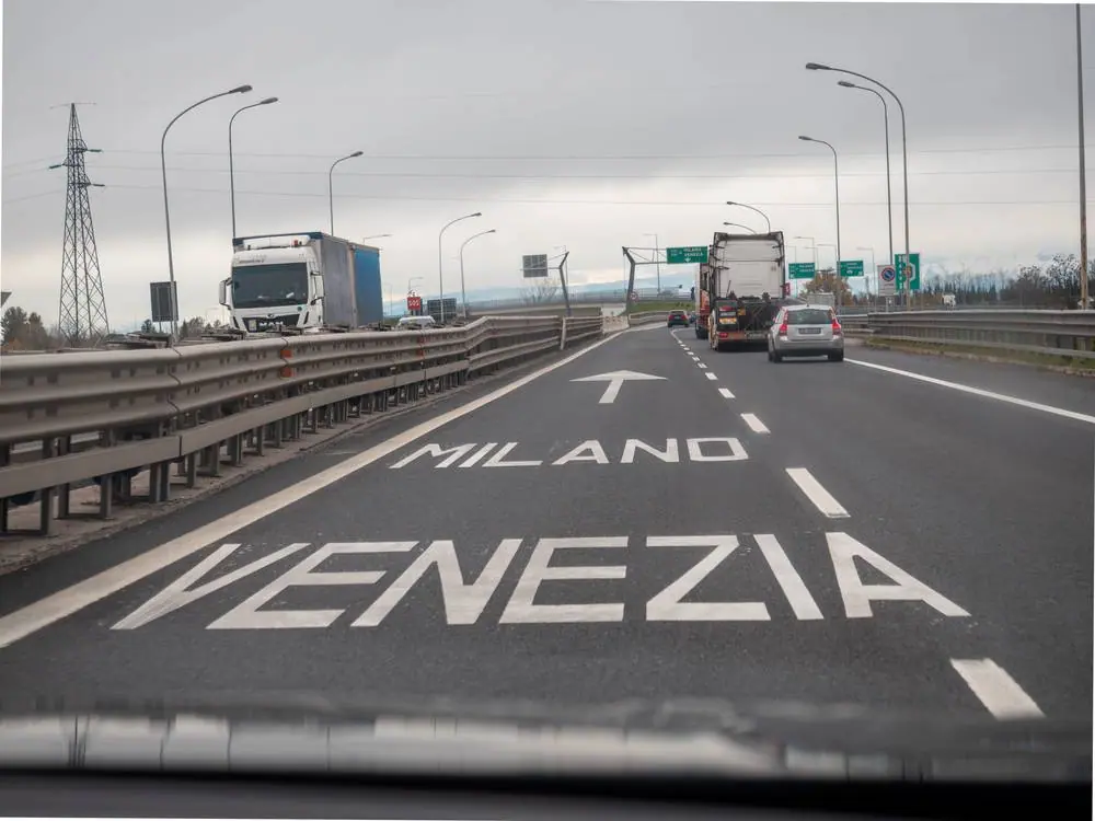 Pohled z auta na italskou dálnici A4 s nápisem Venezia a Milano přímo na vozovce, kamiony a šedé nebe