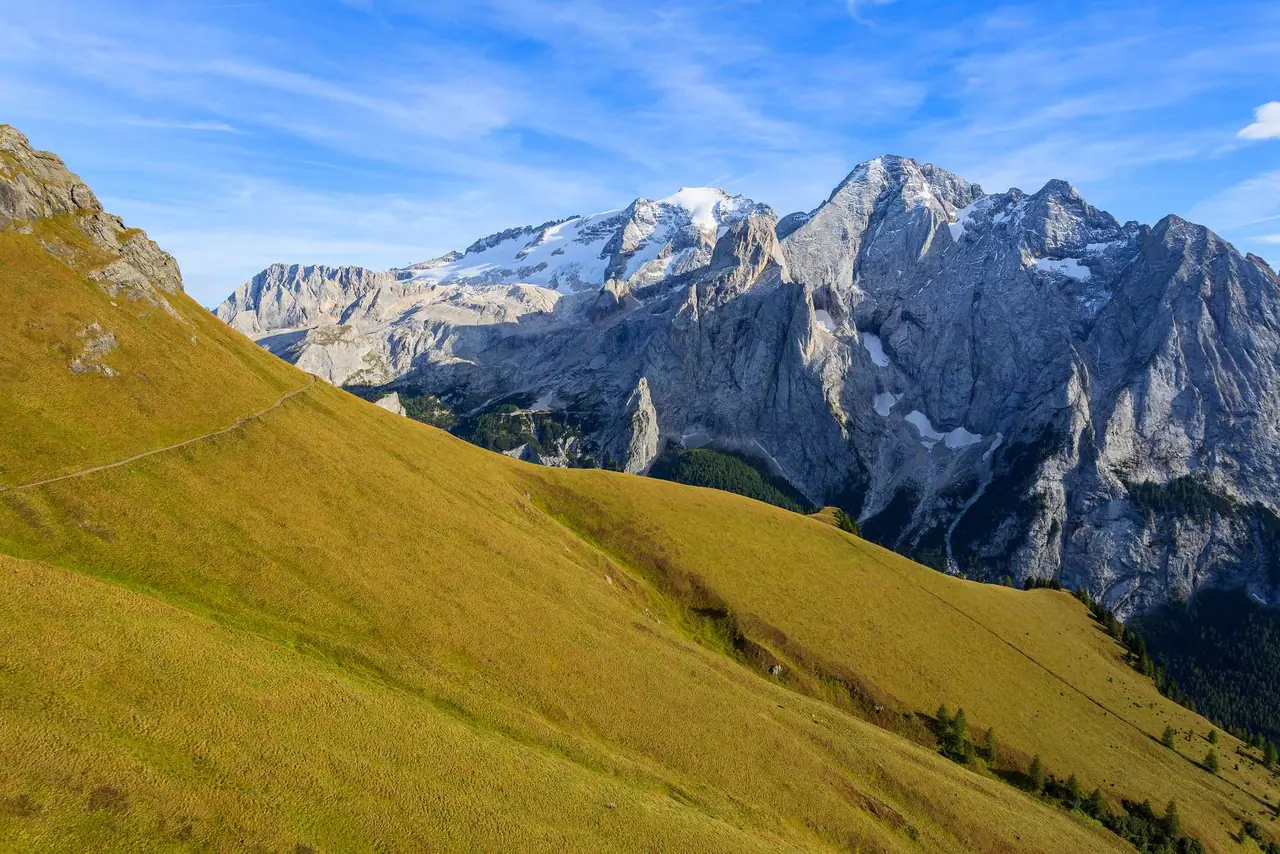 Letní turistická stezka v Dolomitech s výhledem na zasněžené štíty Marmolady a skalnaté útesy nad zeleným svahem
