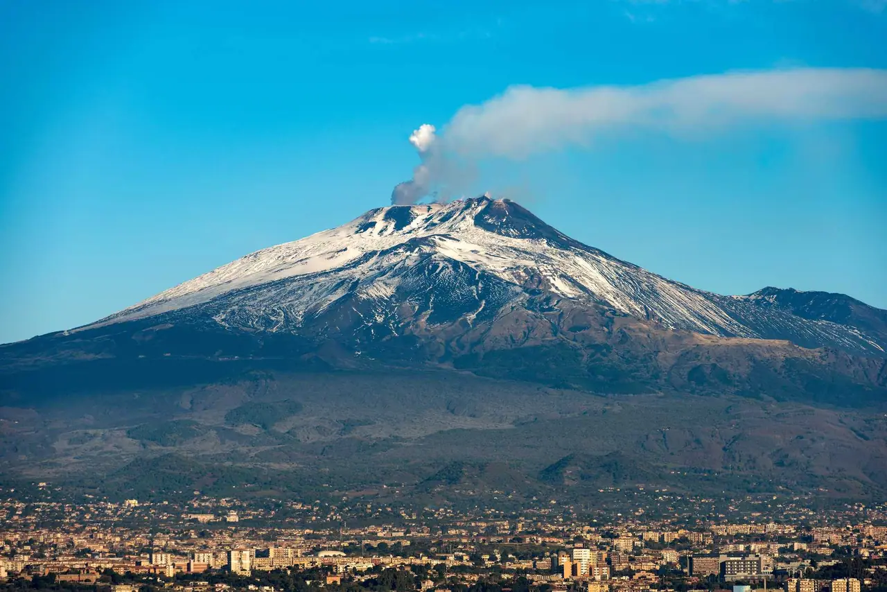 Pohled na aktivní sopku Etna nad Catanií, z vrcholu stoupá sloup kouře a vrchol pokrývá sníh