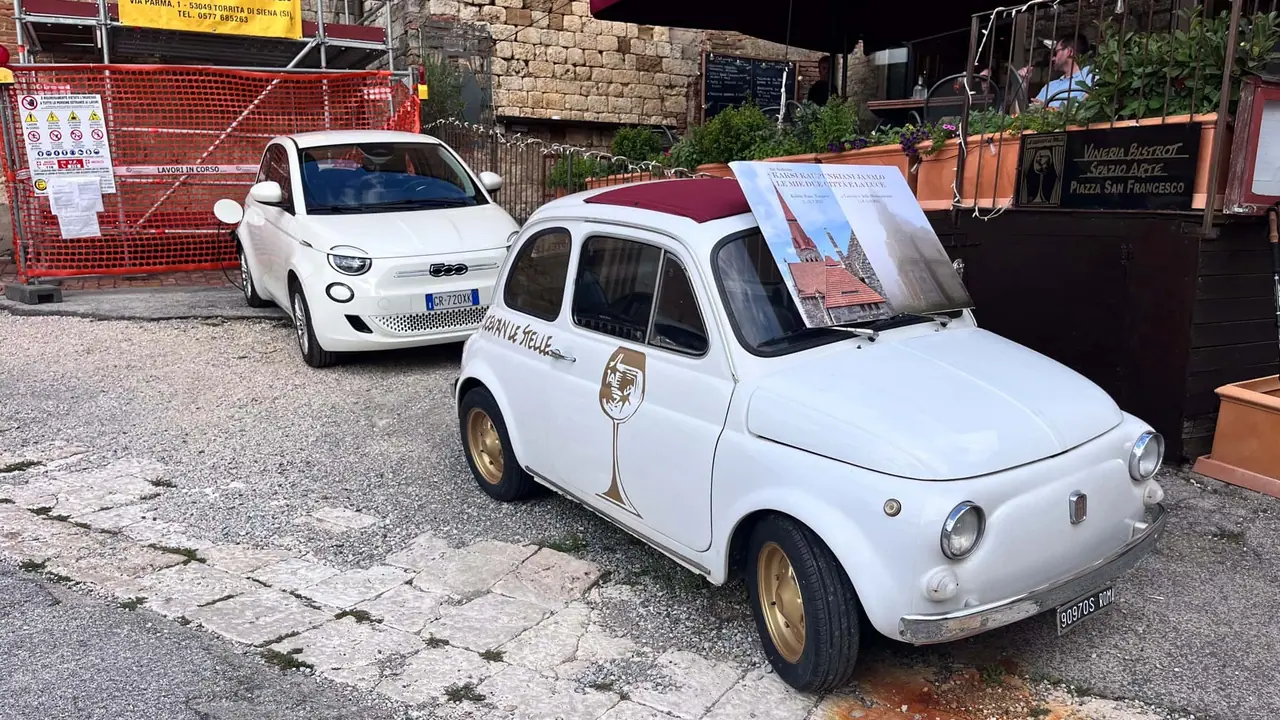 Bílý klasický Fiat 500 a moderní elektromobil zaparkované na dlážděné ulici v Montepulcianu s kamennými zdmi a výkladem restaurace