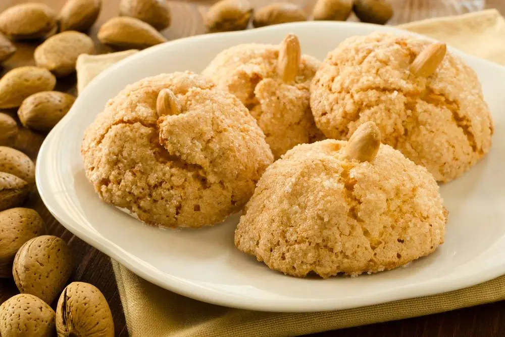 Tradicni italske amaretti susenky s mandlemi na bilem taliri, kolem rozlozene celé mandle v skorapkach na drevenem stole