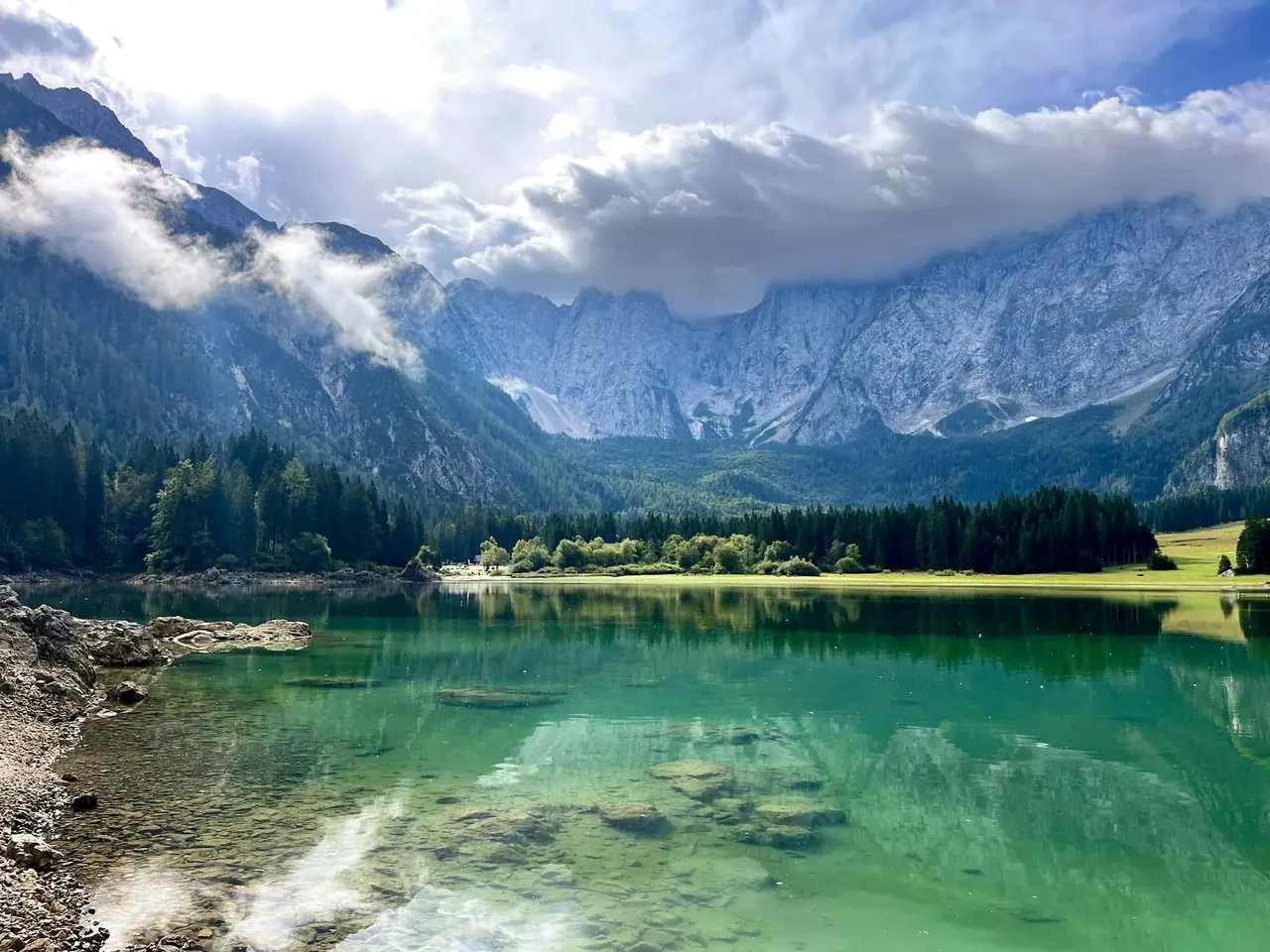 křišťálově čisté jezero Fusine s tyrkysovou vodou obklopené tmavými jehličnany a dramatickými vrcholy Julských Alp s mraky