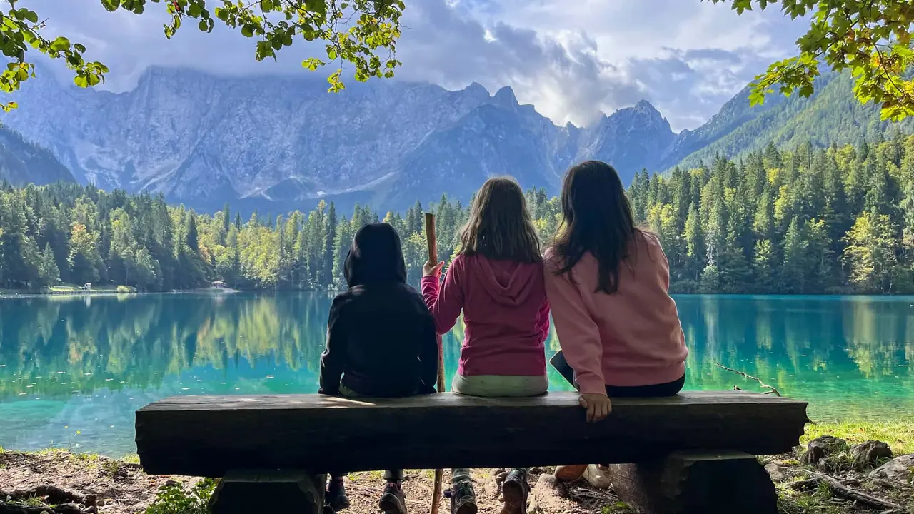 Děti a Monča sedící na lavičce u Lago di Fusine Inferiore sledují horské panorama