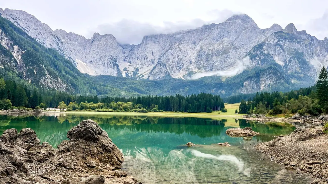 Křišťálově čiré vody Lago di Fusine s balvany na břehu a dramatickými alpskými štíty Julských Alp v pozadí