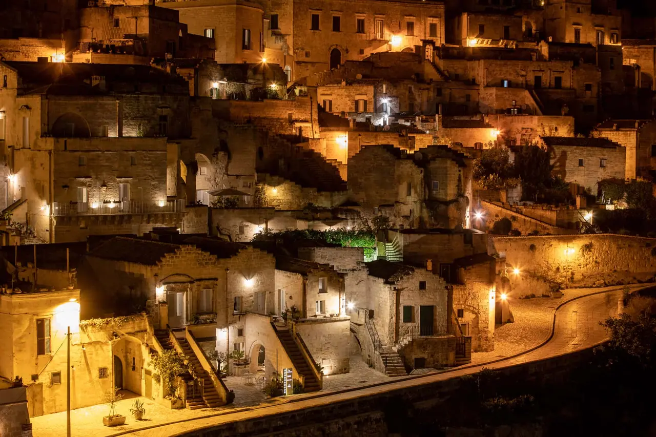 Kouzelný večerní pohled na osvětlené jeskynní město Sassi di Matera s teplým světlem prosvítajícím okny domů po setmění