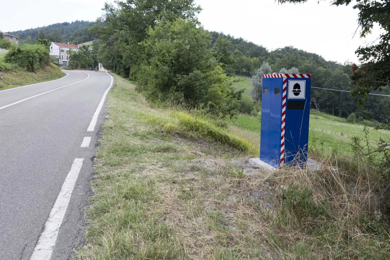 Modrý autovelox stojí u krajnice klikaté silnice mezi zeleným kopci, typický příklad rychlostního radaru který potkáme při cestování Itálií