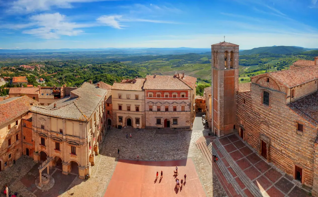 Pohled na hlavní náměstí Piazza Grande v Montepulciano s vysokou věží, kamennými budovami a rodinami procházejícími se po dlažbě