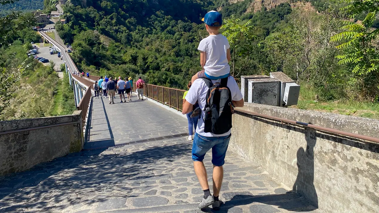 Skupina turistů prochází mostem vedoucím k městu Civita di Bagnoregio, obklopeným zelenými svahy a kaňonem.