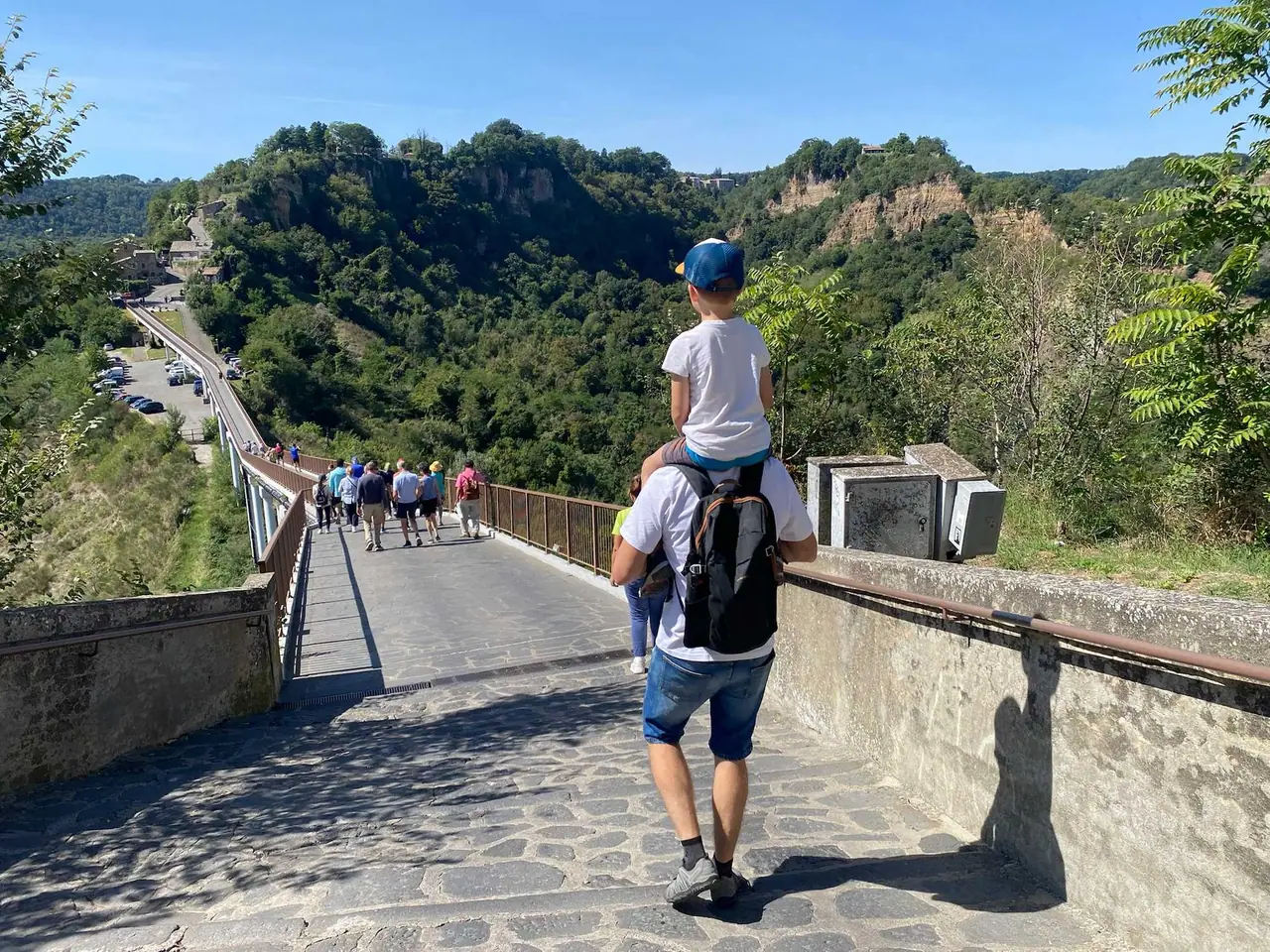 Táta s Filípkem na ramenou kráčí po betonovém mostě vedoucím do Civita di Bagnoregio mezi zelenými kopci