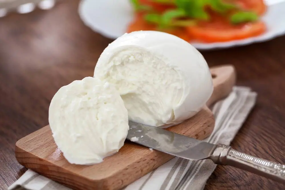 Čerstvá mozzarella nakrájená na dřevěné desce s nožem, v pozadí talíř s rajčaty a bazalkou na dřevěném stole