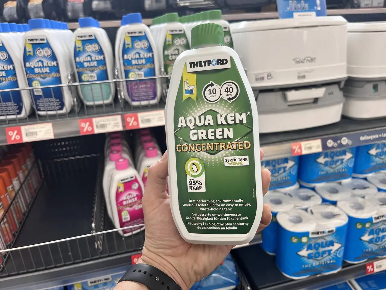 Držíme v ruce zelenou láhev Aqua Kem Green koncentrované toaletní chemie v regálu obchodu mezi dalšími produkty