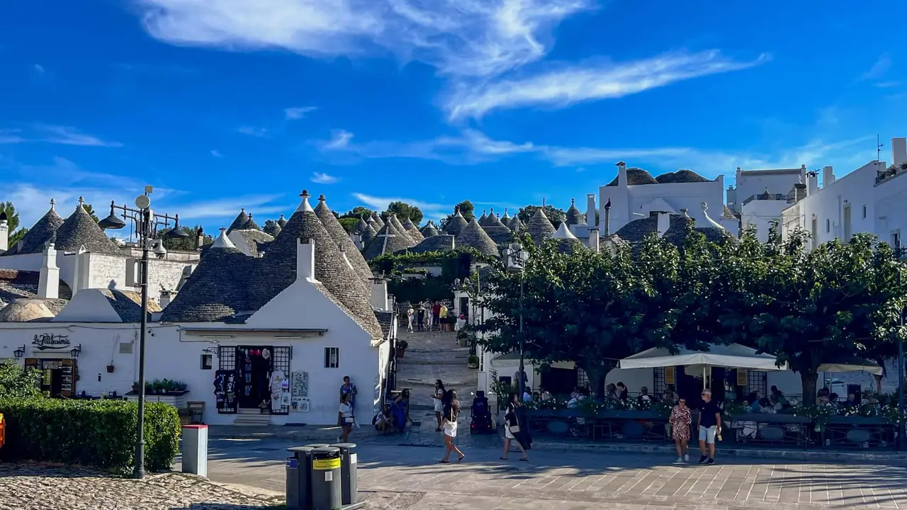 Panoramatický pohled na historické centrum Alberobella s desítkami trulli domečků s kuželovitými střechami, turisté na náměstí a kavárny s terasami