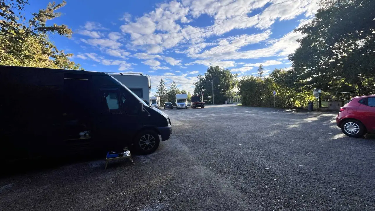 Naše tmavá dodávka zaparkovaná na štěrkovém parkovišti mezi ostatními vozy, kolem stromy a modré nebe s bílými mraky při cestě ze San Marina