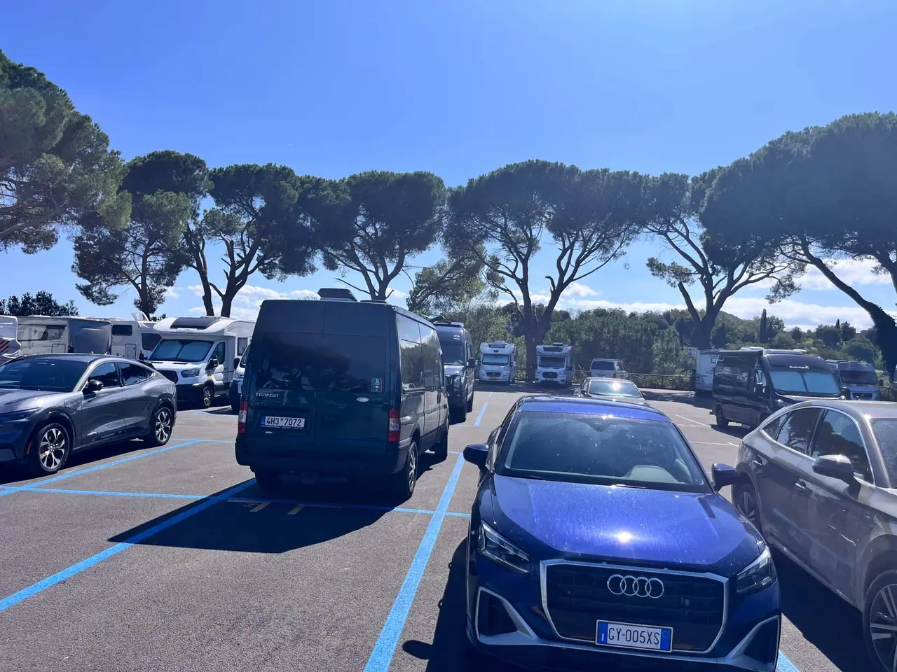 Přeplněné parkoviště v Montepulcianu s obytným vozem, modrou Audi a dalšími auty mezi typickými italskými borovicemi pod jasnou oblohou