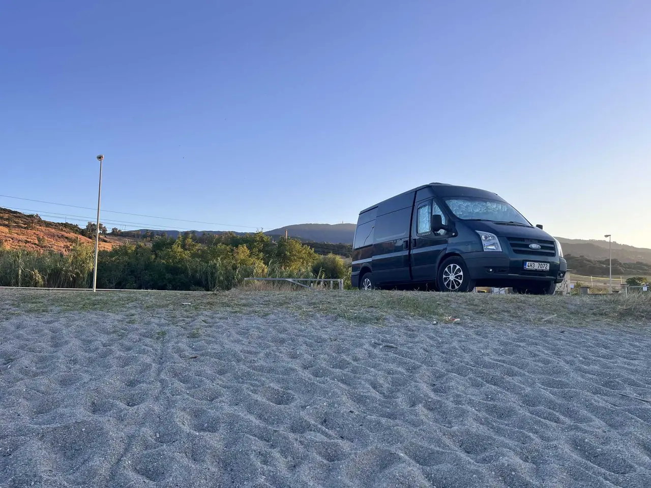 Náš tmavý Ford Transit zaparkovaný na písčitém parkovišti u pláže za Falerna Scalo s kopci v pozadí při večerním světle
