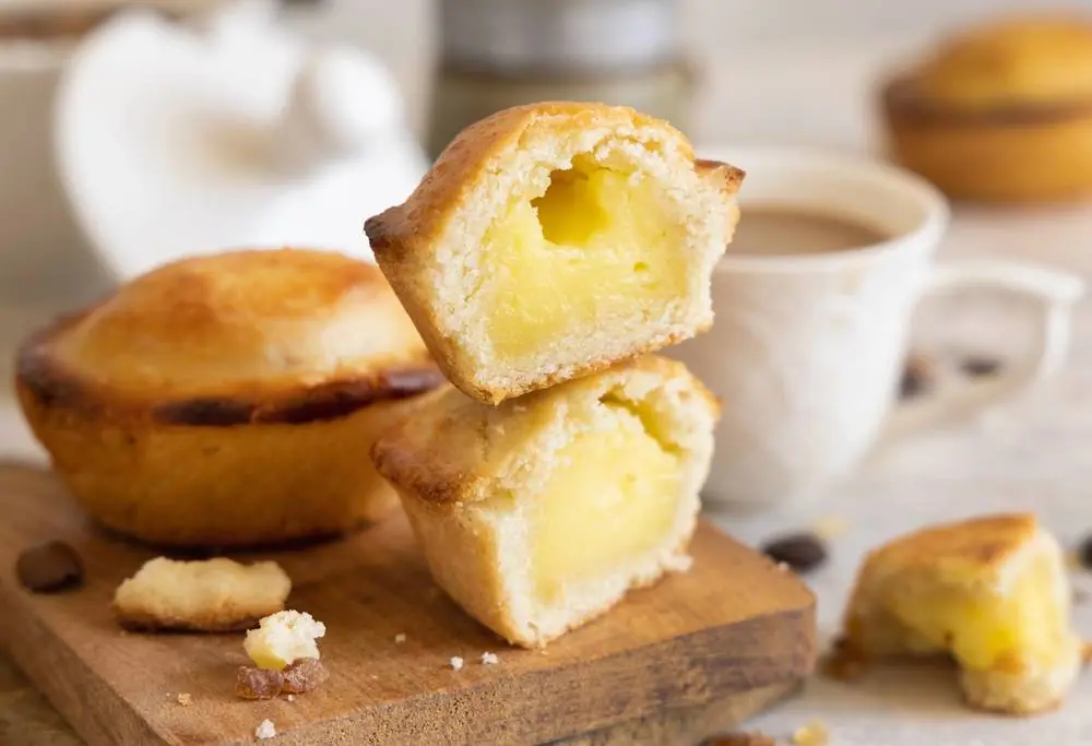 Čerstvě upečené pastéis de nata s vanilkovým krémem na dřevěném prkénku, jeden rozkrojený ukazuje zlatavou náplň, vedle káva