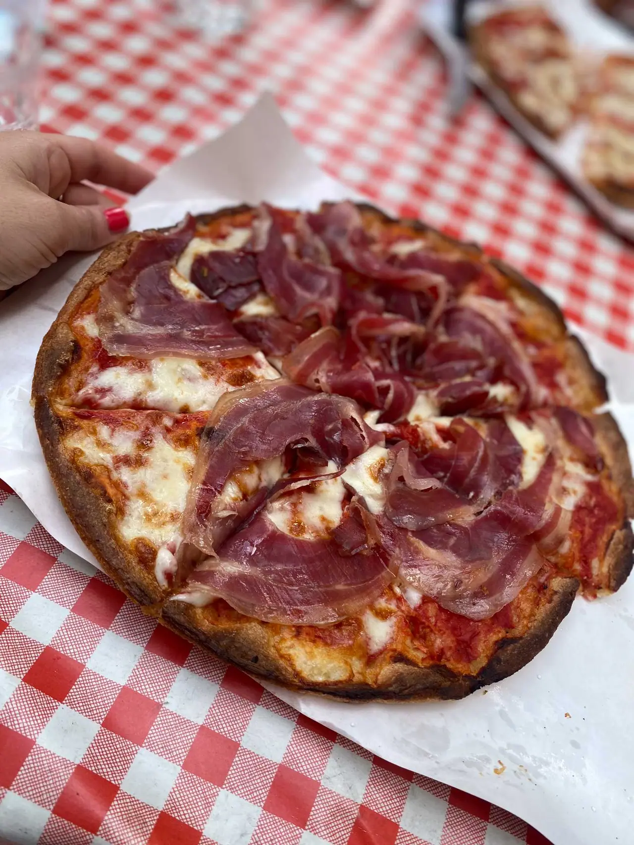 Naše čerstvá pizza s italským pršutem na červenobílém ubrousku v pizzerii ve Fivizzanu, ruka sahá po kousku