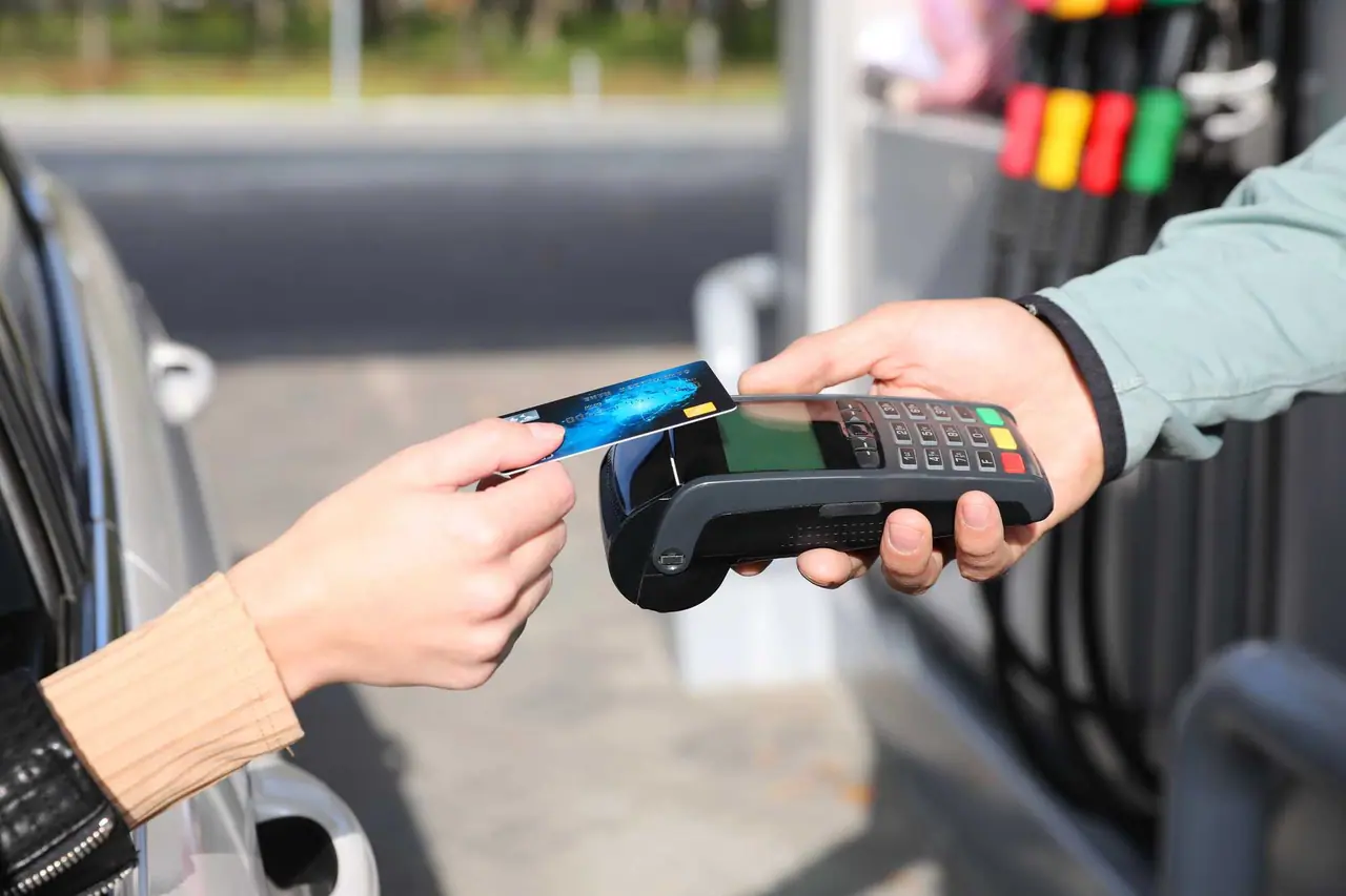 Ruka přikládá modrou platební kartu k terminálu, který drží obsluha čerpací stanice s farebnými pistolemi v pozadí