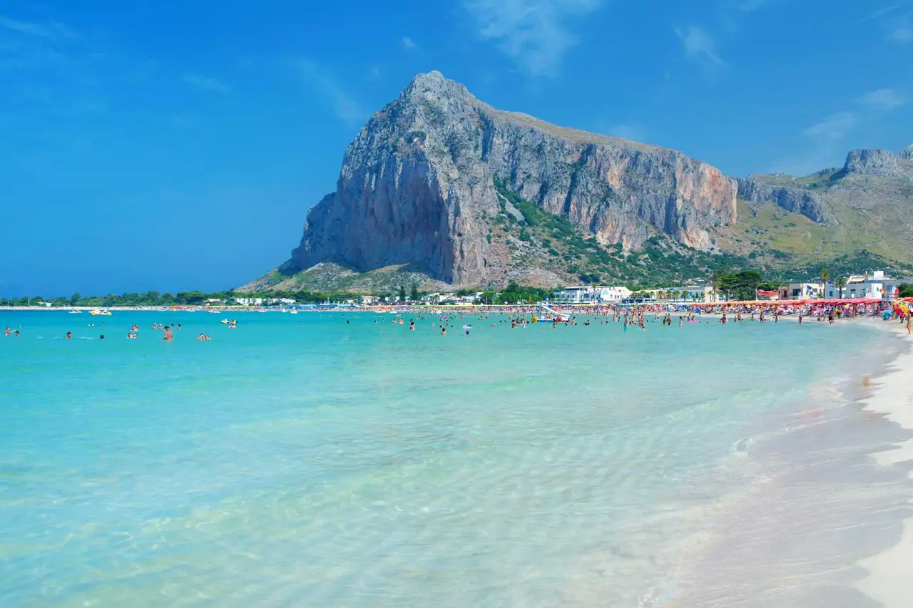 Průzračné tyrkysové moře pláže San Vito lo Capo na Sicílii s masivem Monte Monaco v pozadí, plné koupajících se lidí