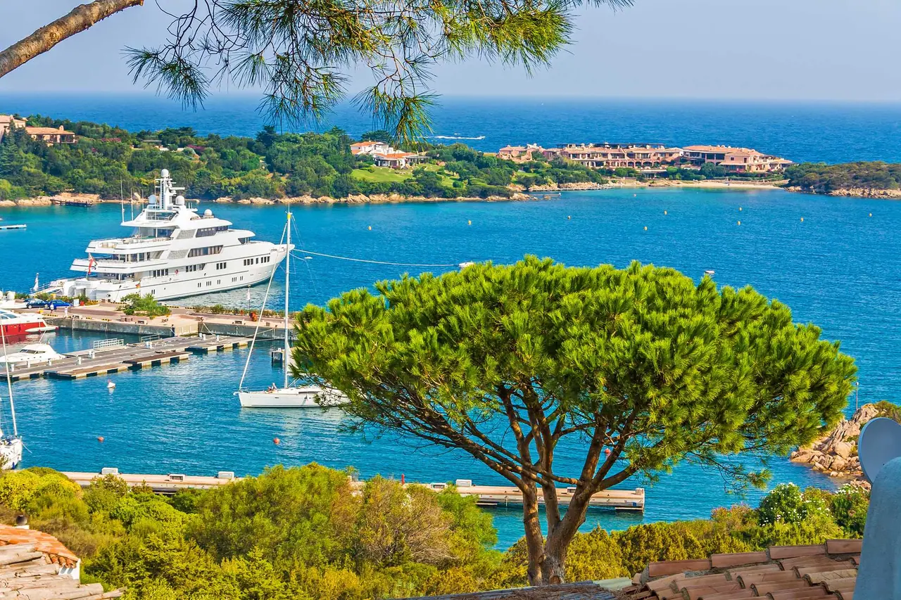 Pohled na přístav Porto Cervo na Sardinii s obří bílou jachtou, tyrkysovým mořem a středomořskými borovicemi v popředí