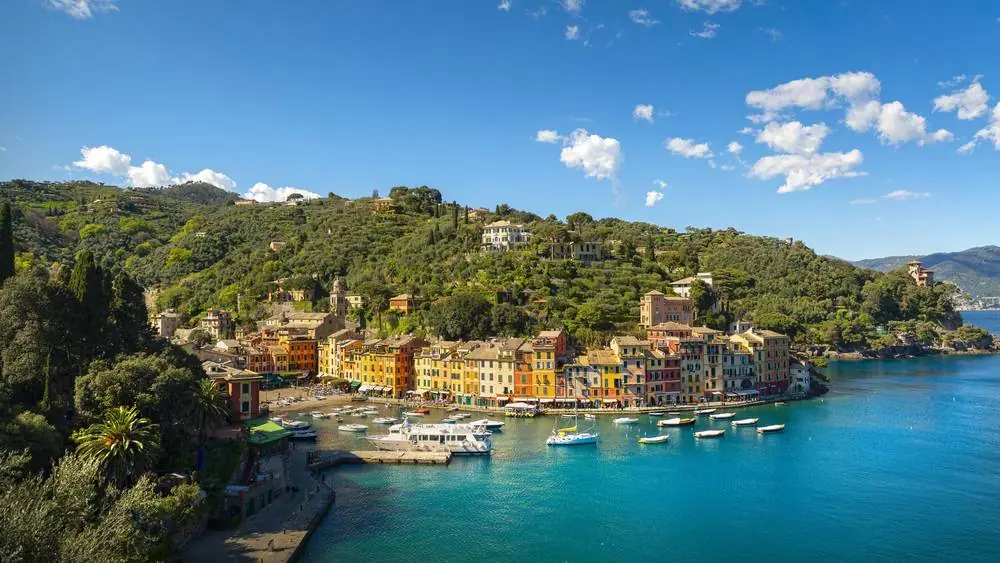 Portofino přístav s jachtami na tyrkysové vodě, barevné italské domy oranžové a žluté podél zálivu, zelené zalesněné kopce v pozadí