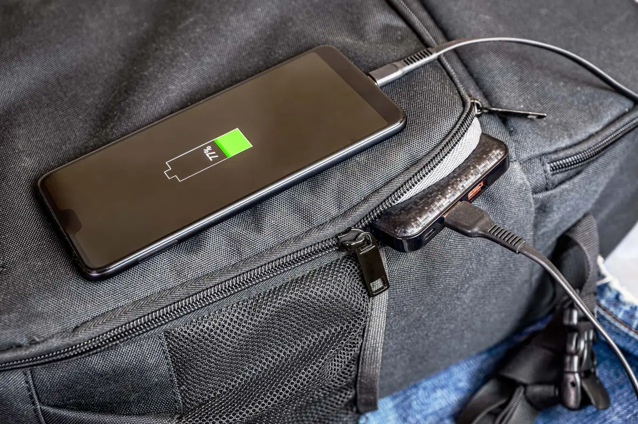 Mobilní telefon nabíjený přes powerbanku na batohu – nezbytná elektronika na cesty.