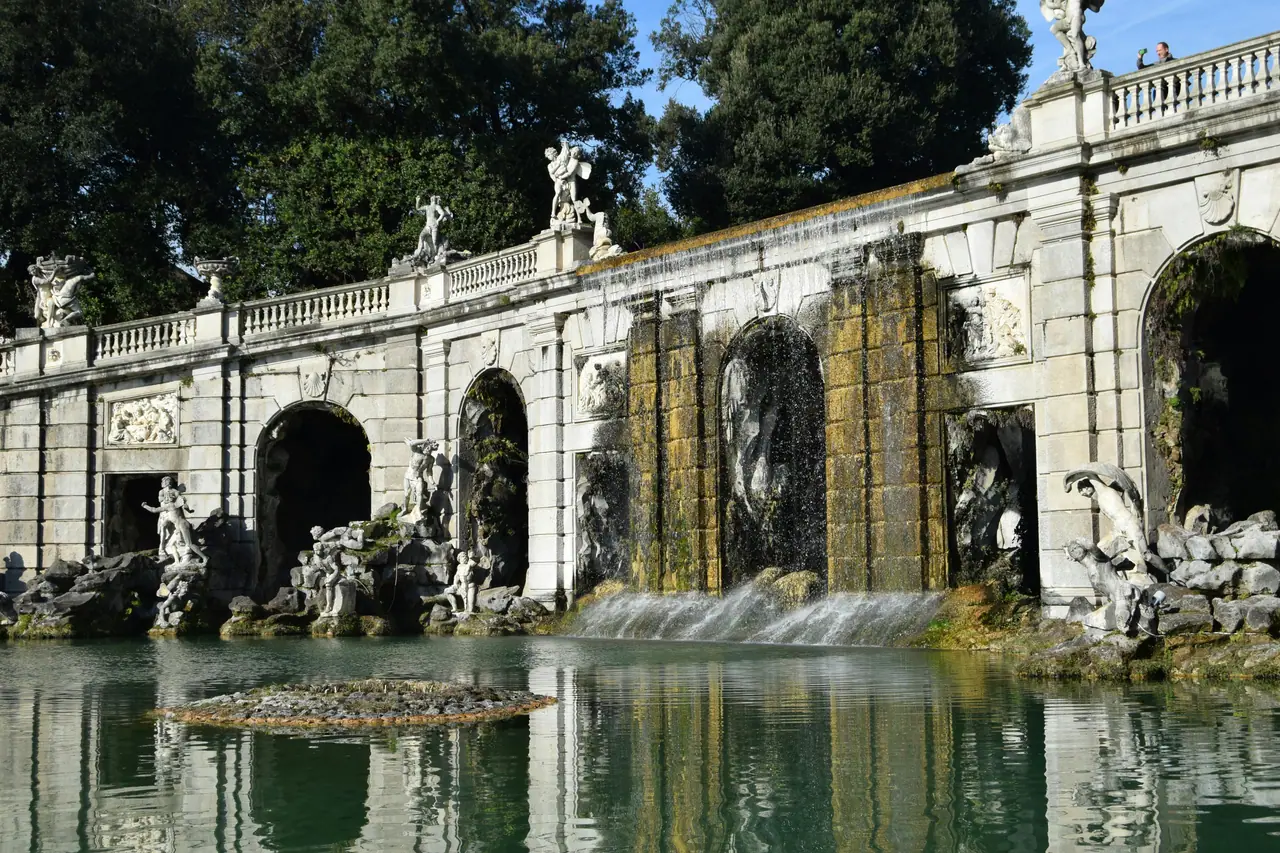 A magnificFontána Aiolos (Fontana di Eolo) je jednou z nejpůsobivějších fontán v Královském paláci v Casertě. Je zasvěcena Aiolovi, mytologickému bohu větrů, který měl moc ovládat vzdušné proudy. ent Baroque-style fountain with sculptures in a tranquil park setting, reflecting elegance and nature.