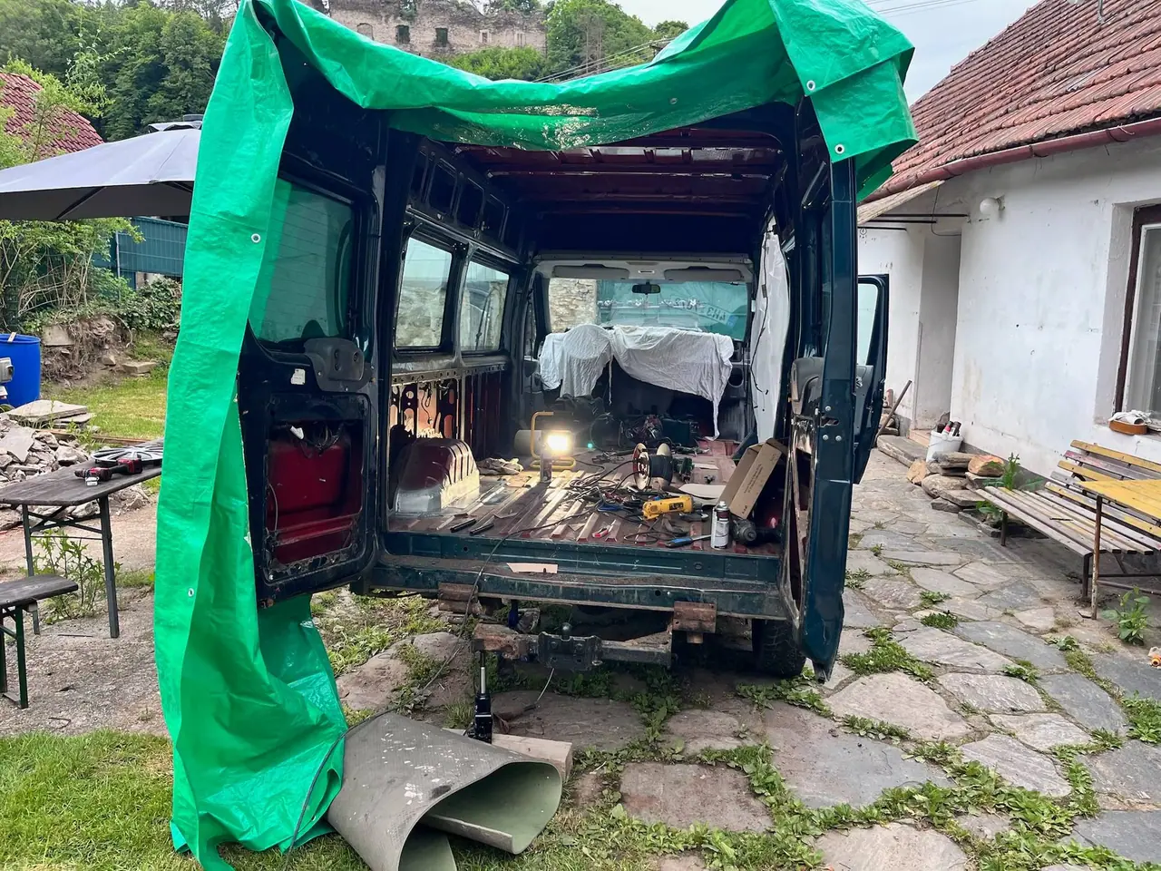 Naše rozložená dodávka Transit pod zelenou plachtou během renovace, s nástroji a materiálem rozházeným uvnitř na dvoře