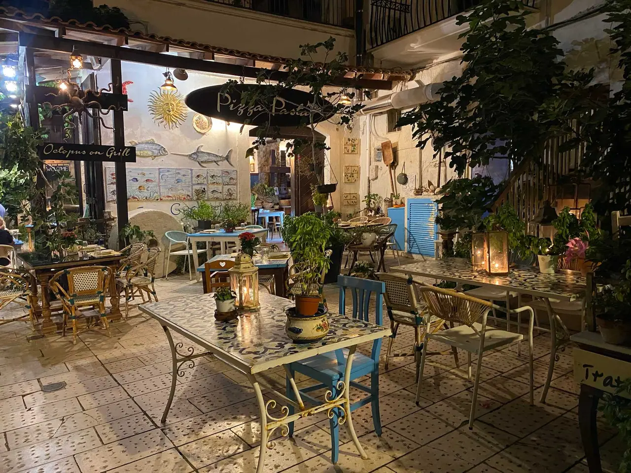Večerní terasa restaurace Pizza Pasta ve Vieste s osvětlenými stolky, květinami a tyrkysovými židlemi pod balkónem