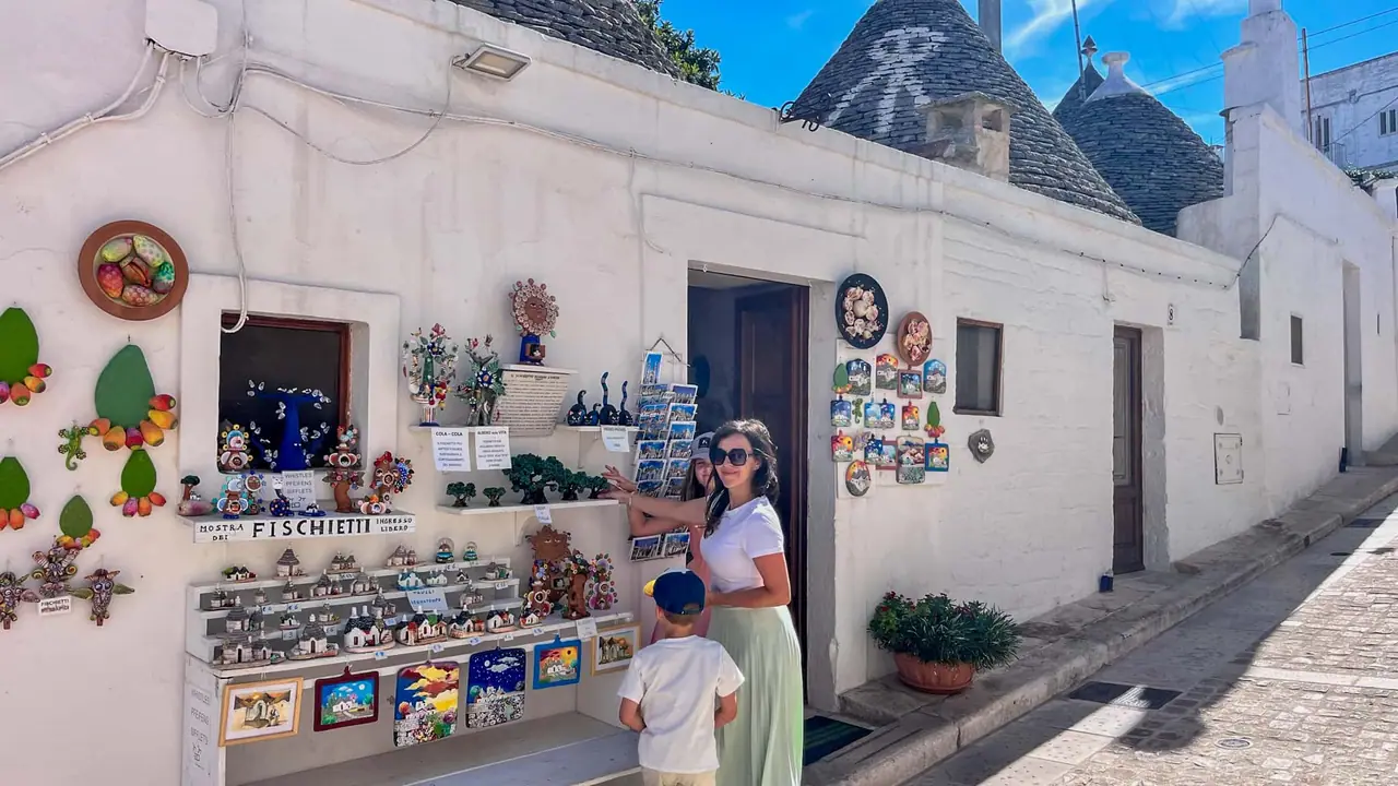 Monča s dětmi u obchodu Fischietti s tradičními suvenýry a řemeslnými výrobky v bílém trullo domečku v Alberobellu