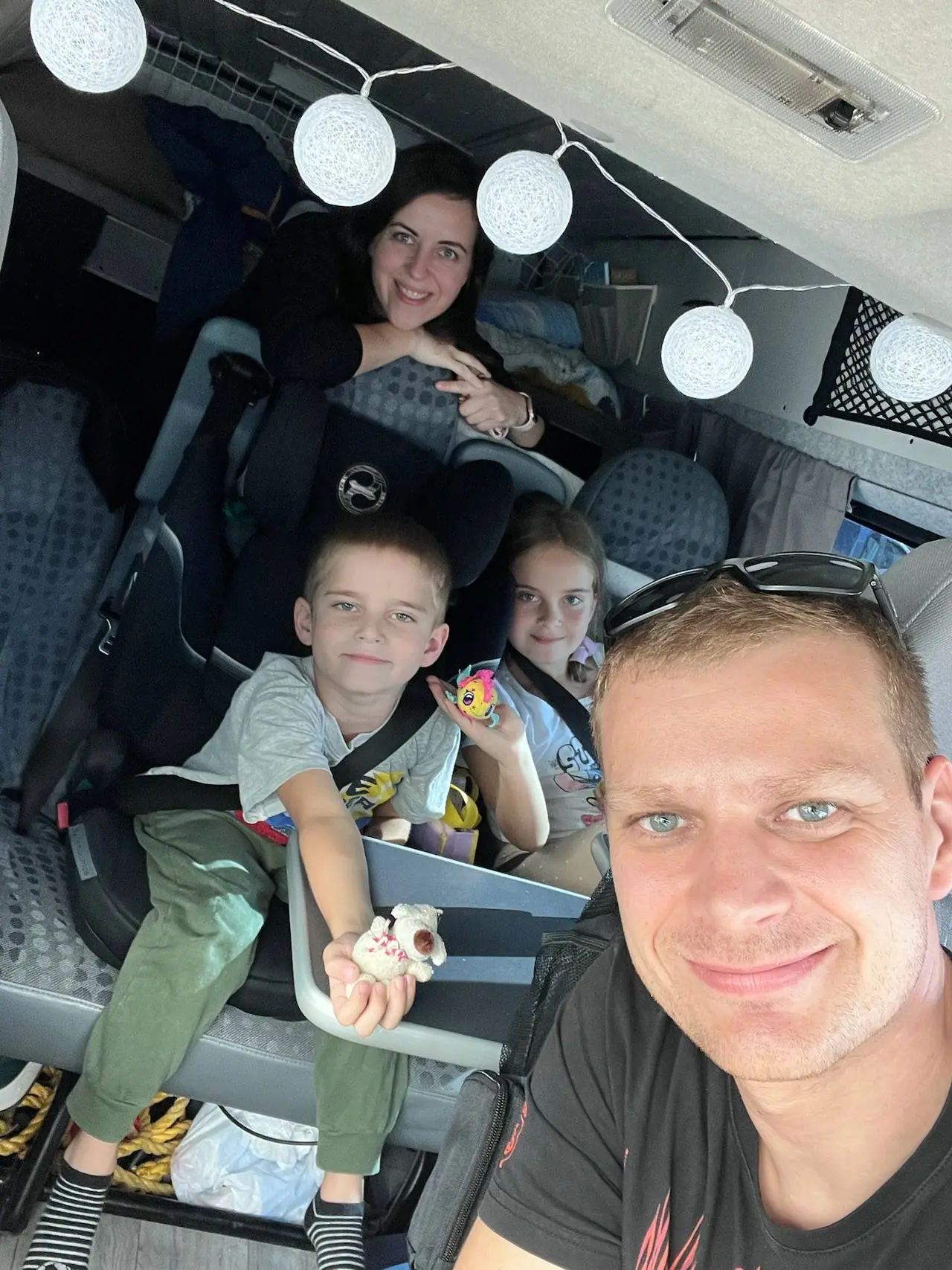 Selfie celé naší rodiny uvnitř karavanu první den cesty - táta v popředí, za ním syn a dcera s hračkami, maminka vzadu mezi ozdobnými světýlky
