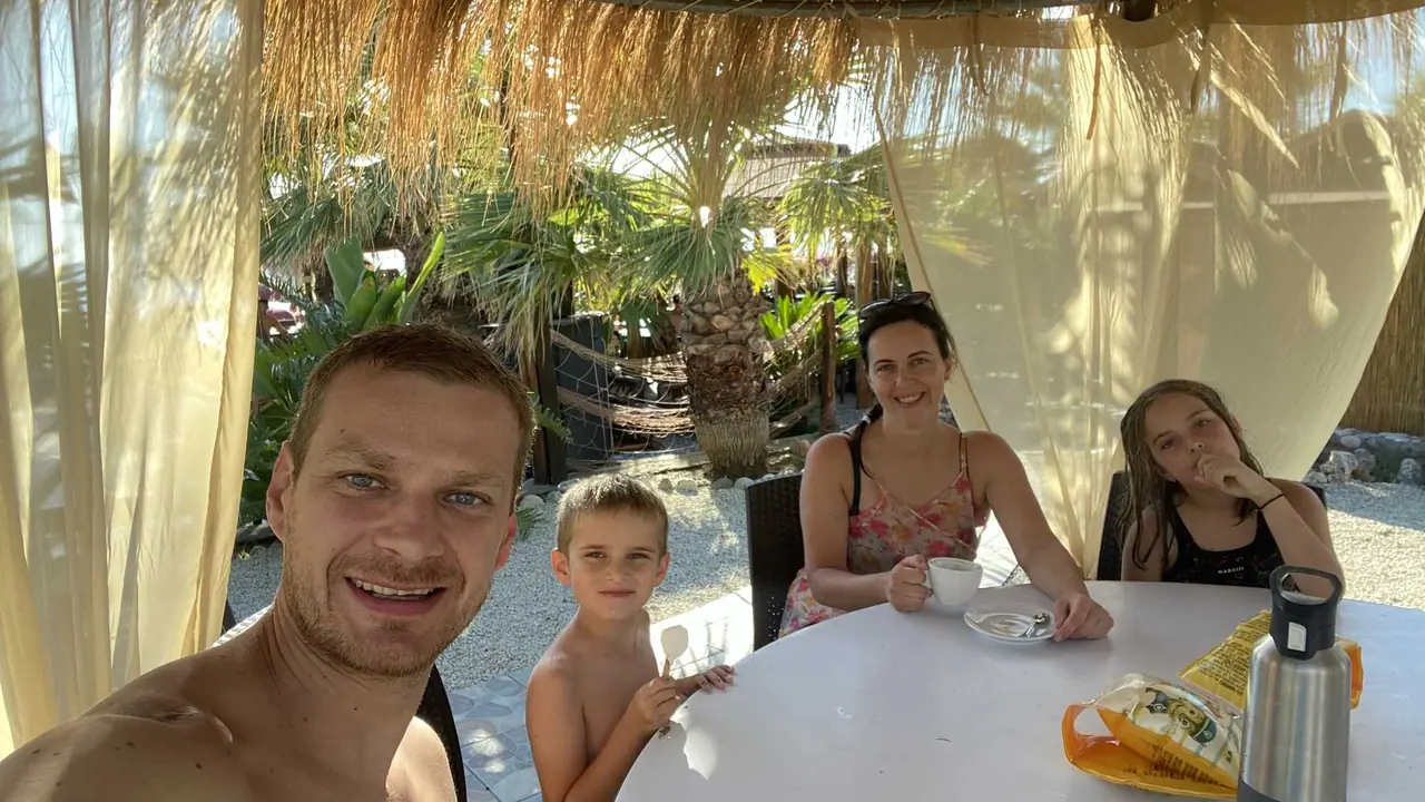 Rodinná siesta pod slunečníkem v Lido Palm Beach Clubu - otec s dítětem a dvě ženy u bílého stolku mezi palmami