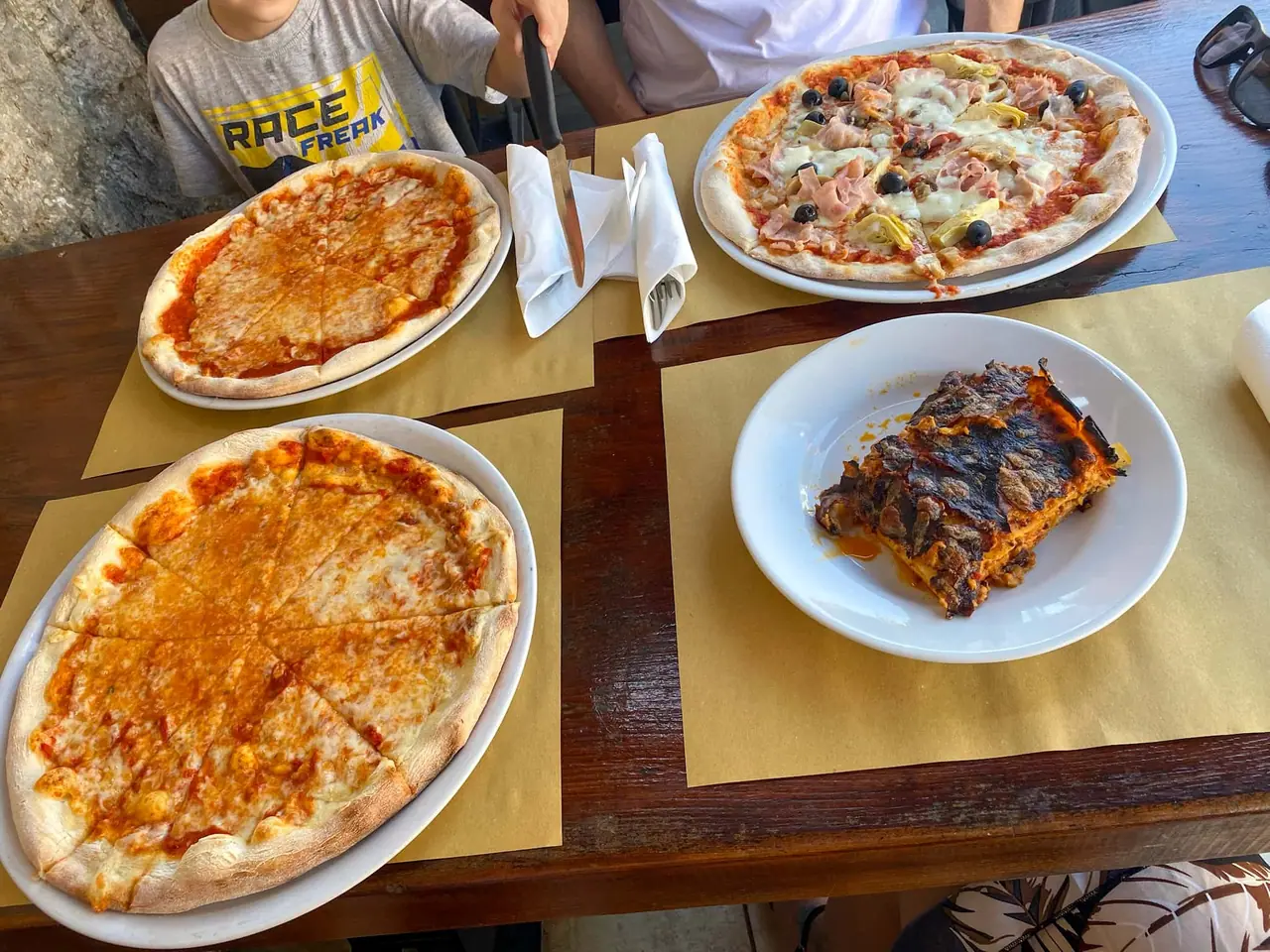 Náš rodinný oběd v Montepulcianu - tři pizzy a lasagne na dřevěném stole, kde jsme si pochutnávali na místní kuchyni