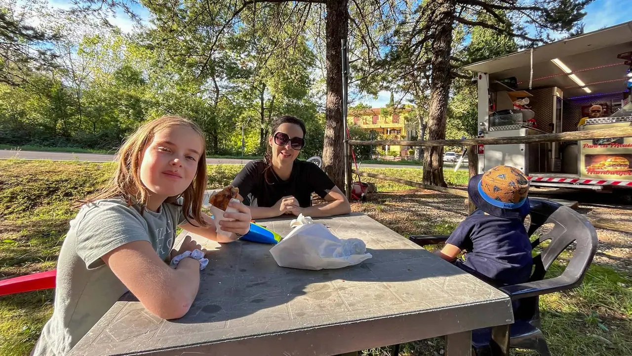 Naše rodinná pauza u food trucku v Toskánsku - sedíme u dřevěného stolu pod stromy a pochutnáváme si na jídle.