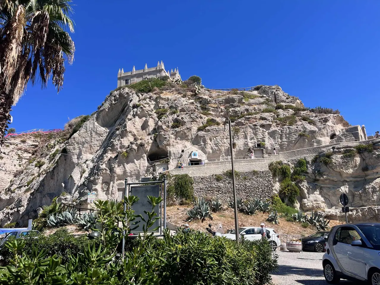 Bílý kostel Santa Maria dell'Isola na skalnatém útesu v Tropei s modrou oblohou, palmami a parkujícími auty