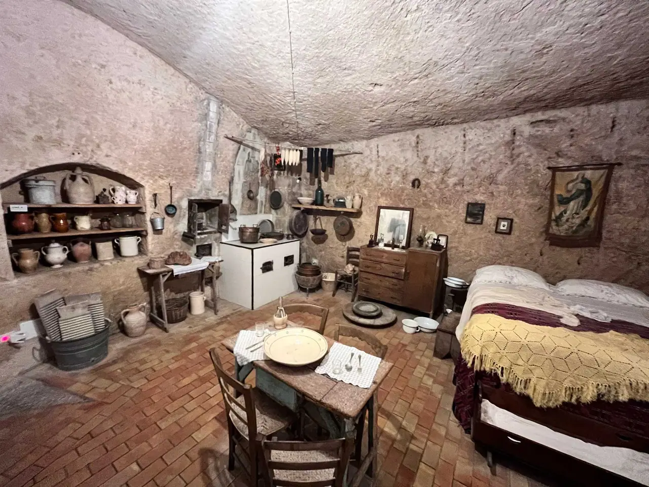 Autentický interiér jeskynního obydlí v Sassi di Matera s cihlovou podlahou, kamennými zdmi, postelí s barevnou dekou a historickým nábytkem