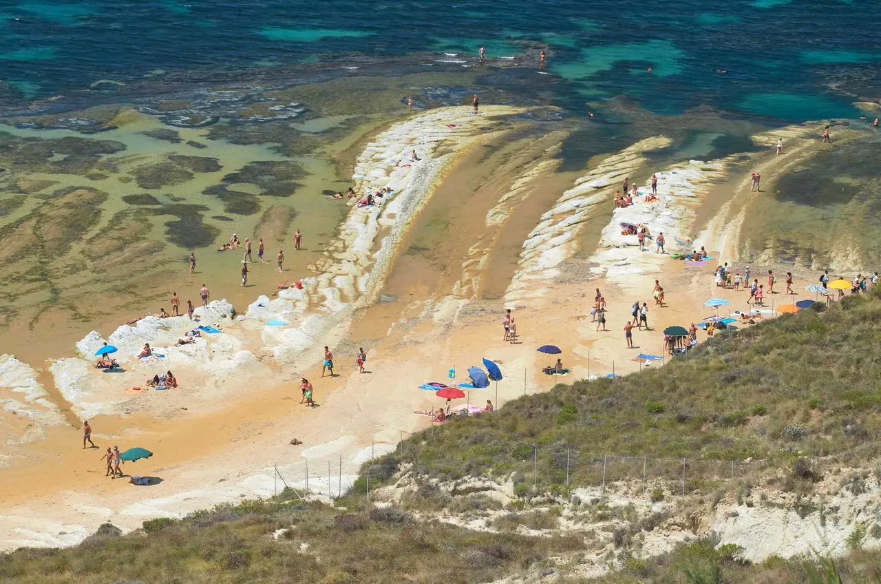 Pohled shora na bílé vrstevnaté skály Scala dei Turchi na Sicílii, kde turisté relaxují u tyrkysového moře s barevnými slunečníky.