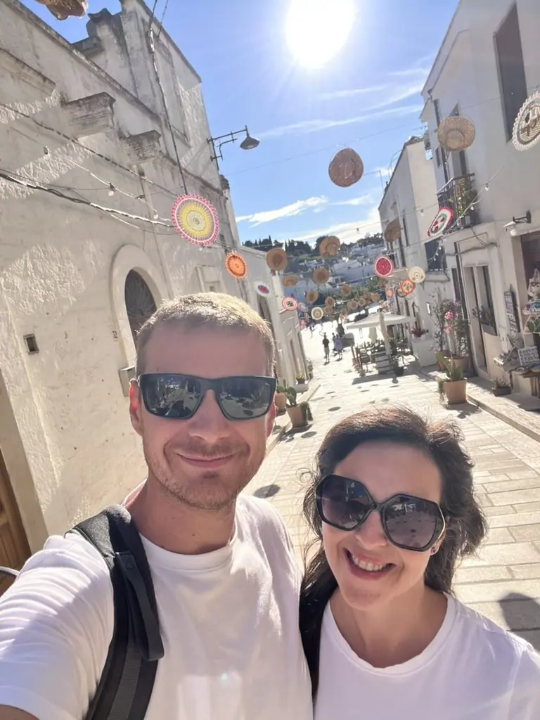 Naše selfie na dlážděné ulici Strada dei Cappelli v Alberobello s klobouky visícími nad hlavami mezi bílými trulli domky